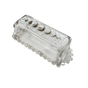 May include: A clear glass butter dish with a rectangular shape. The lid has a series of round, raised glass accents. The base features a beaded edge. The dish is designed to hold a standard size of butter.