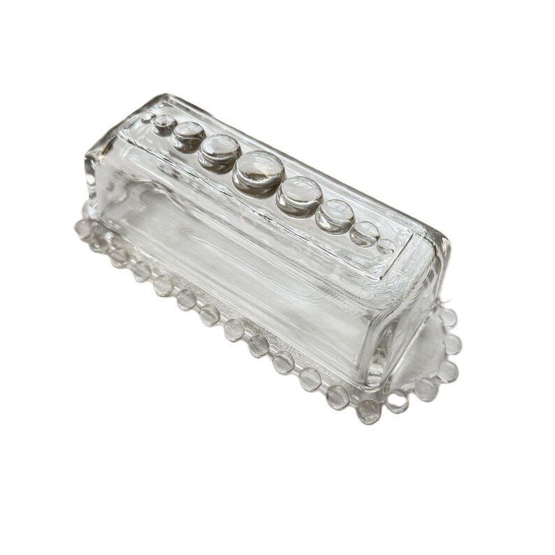May include: A clear glass butter dish with a rectangular shape. The lid has a series of round, raised glass accents. The base features a beaded edge. The dish is designed to hold a standard size of butter.