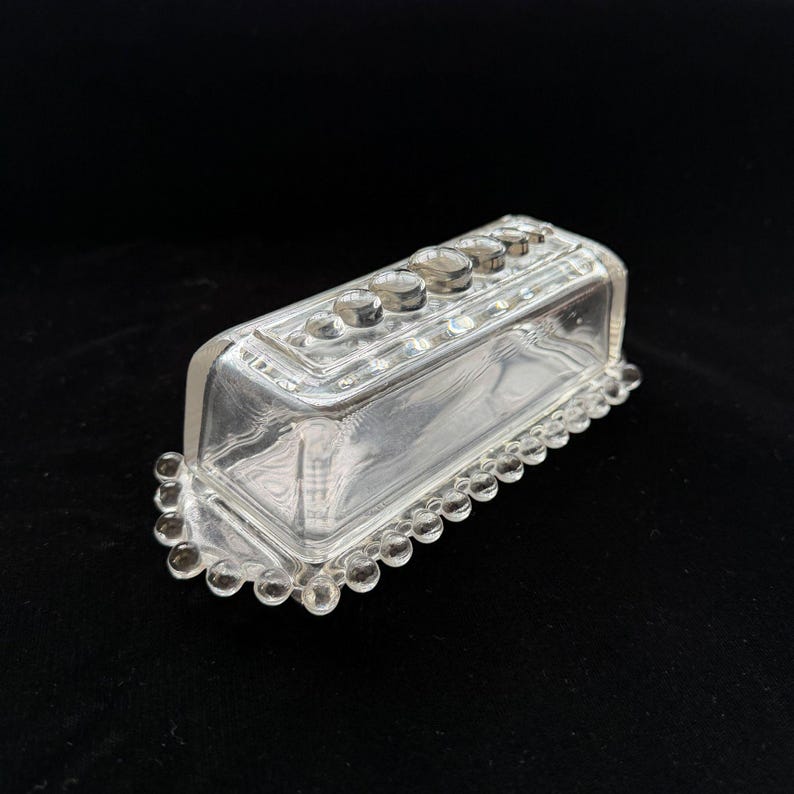 May include: A clear, rectangular glass butter dish with a domed lid. The lid has a row of small, round knobs. The base features a beaded edge. The dish is designed to hold a standard size of butter.