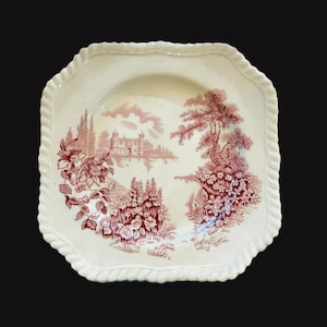 May include: Square, off-white plate with a red and white landscape design. The plate features a detailed illustration of a building, trees, and flowers. The rim has a textured, rope-like border.