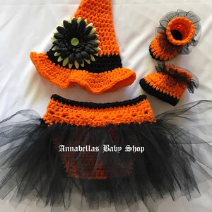 May include: A crocheted witch costume for a baby, featuring a black and orange hat with a flower, a black and orange tutu, and matching booties.