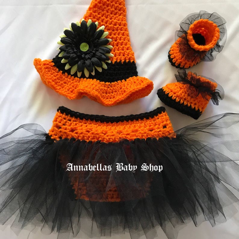 May include: A crocheted witch costume for a baby, featuring a black and orange hat with a flower, a black and orange tutu, and matching booties.