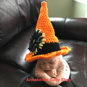 May include: A crocheted witch hat for a baby, with an orange and black color scheme. The hat has a black flower with green accents on the brim. The hat is on a baby lying on a brown leather couch. The text "Annabellas Baby Shop" is visible at the bottom of the image.
