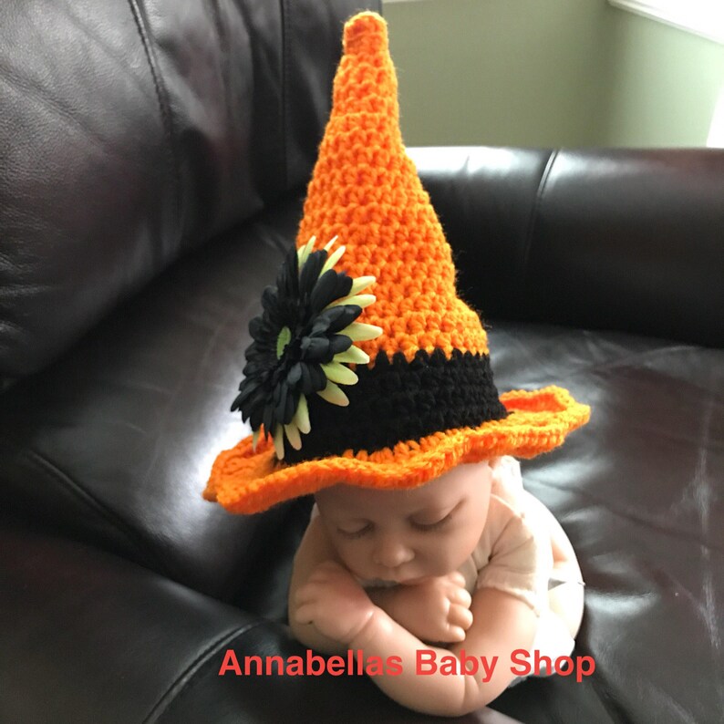 May include: A crocheted witch hat for a baby, with an orange and black color scheme. The hat has a black flower with green accents on the brim. The hat is on a baby lying on a brown leather couch. The text "Annabellas Baby Shop" is visible at the bottom of the image.