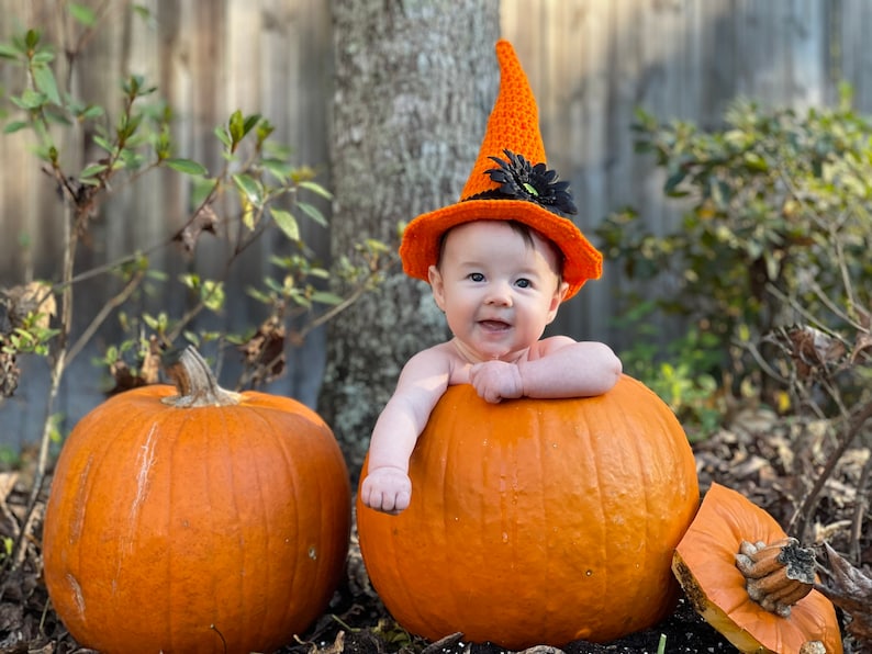 May include: A baby wearing an orange crocheted witch hat with a black flower sits inside a large orange pumpkin. A smaller orange pumpkin is on the left side of the image.