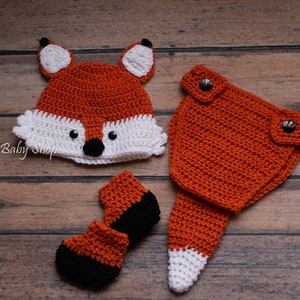 May include: A crocheted fox costume for a baby, featuring a hat, pants, and booties. The hat has a white face with black eyes and nose. The pants have a tail and the booties have black accents.