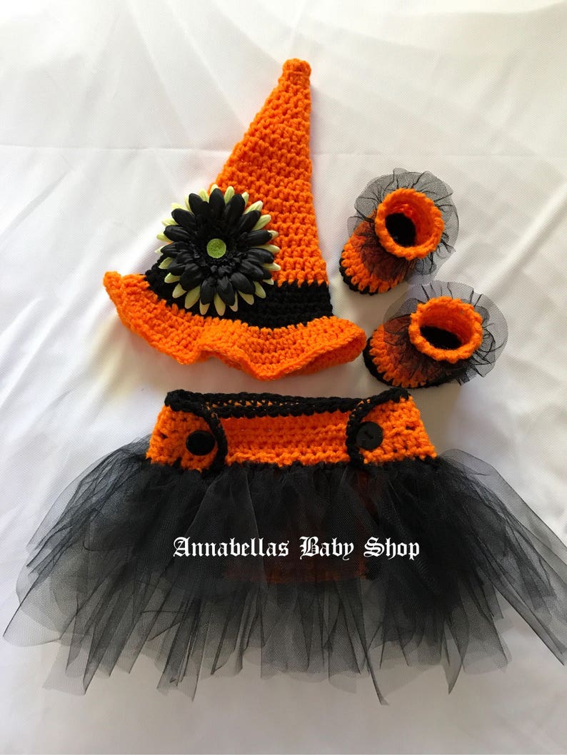 May include: A crocheted witch costume for a baby, featuring a black and orange hat with a flower, a black and orange tutu, and matching booties.