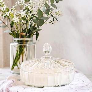 May include: A clear, lidded glass jar with a ribbed design, filled with a white substance, sits on a white lace cloth. A glass vase with white flowers and green foliage is in the background, creating a serene, decorative scene.