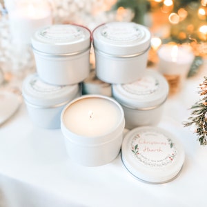 May include: White soy wax candles in small, round tins. One candle is lit, with a visible flame. The lids feature a wreath design and the words "Christmas Hearth". The candles are arranged on a white surface, with a blurred background.