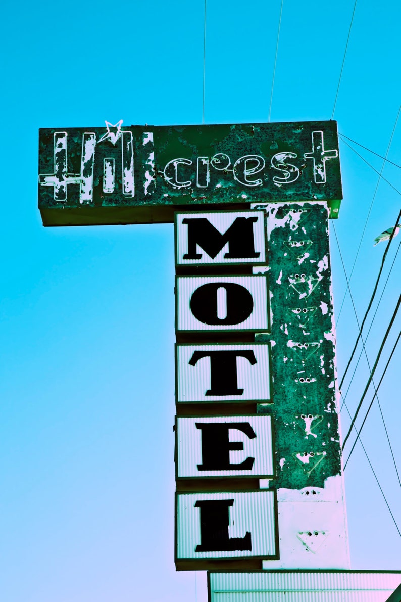 May include: A vintage motel sign with faded green paint and white lettering. The sign reads "Hillcrest Motel" with the word "Motel" stacked vertically in black letters.