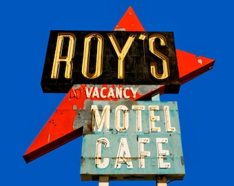 May include: A vintage motel sign with a red and white star shape. The sign reads "Roy's Motel Cafe" in gold lettering on a blue background. The top of the sign reads "Roy's" in gold lettering on a black background.
