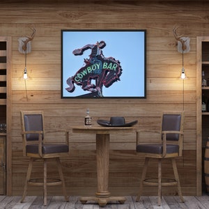 May include: A rustic bar scene with wooden walls and furniture. A framed sign reading "COWBOY BAR" hangs above a small table with two bar stools. Wine racks and a barrel wine cooler are visible.