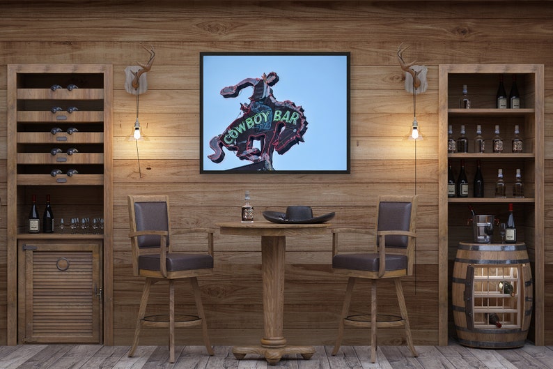 May include: A rustic bar scene with wooden walls and furniture. A framed sign reading "COWBOY BAR" hangs above a small table with two bar stools. Wine racks and a barrel wine cooler are visible.