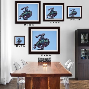 May include: Framed prints of the Cowboy Bar sign, featuring a neon cowboy riding a bucking bronco. The sign, with the words "COWBOY BAR", is set against a blue sky. Various sizes are displayed, including 24 x 20 inches, 30 x 24 inches, and 14 x 11 inches.