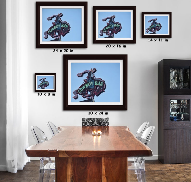 May include: Framed prints of the Cowboy Bar sign, featuring a neon cowboy riding a bucking bronco. The sign, with the words "COWBOY BAR", is set against a blue sky. Various sizes are displayed, including 24 x 20 inches, 30 x 24 inches, and 14 x 11 inches.