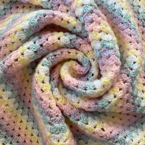 May include: A handmade crochet blanket in pastel colors. The blanket features a granny square pattern in shades of pink, yellow, blue, and lavender. The soft, textured blanket is folded in a spiral, showcasing the intricate stitch work.
