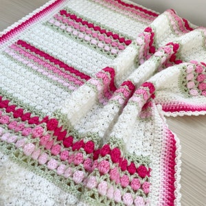 May include: A handmade crochet blanket in white, pink, and green. The blanket features stripes and textured patterns in varying shades of pink and green, with a solid pink border. The soft, textured blanket is folded, showcasing its design.