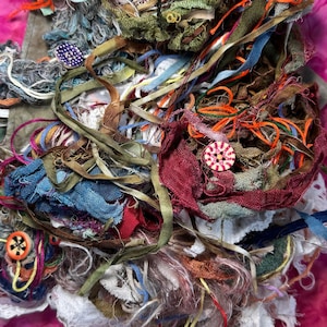 May include: A collection of assorted fabric strips, ribbons, and yarn in various textures and colors including blue, green, red, orange, and white. Two round buttons are visible, one purple with white dots and one white with red stripes.