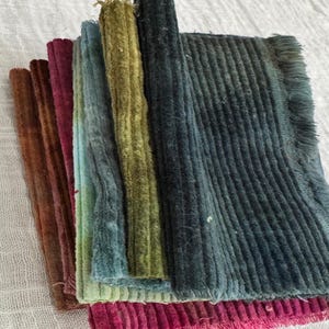 May include: A collection of colorful corduroy fabric samples in shades of brown, burgundy, teal, green, and blue. The fabric is arranged in a layered display, showcasing the texture and color variations. The corduroy has a ribbed texture.