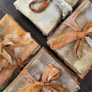 May include: Close-up of several square fabric napkins in shades of beige, brown, and orange, tied with matching frayed ribbon. The napkins have a rustic, handmade appearance, with visible texture and raw edges. The arrangement suggests a set of cloth napkins.