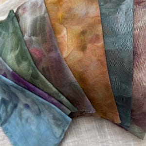 May include: A collection of hand-dyed fabric swatches in various colors, including blue, green, purple, and orange. The fabric has a textured appearance and a watercolor-like effect, with visible brushstrokes and color variations. The edges of the fabric are frayed.