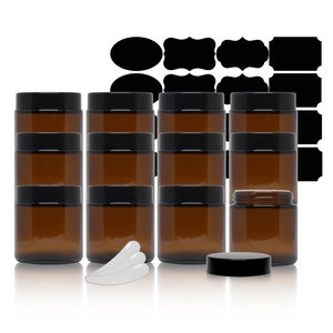 May include: A collection of amber glass jars with black lids, arranged in rows. Several black label stickers of various shapes are in the background. White plastic spatulas are also included.