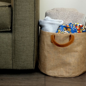 Peut inclure: Un panier de rangement en toile de jute brune avec une poignée en cuir, rempli de serviettes et de tissus pliés. Le panier est placé à côté d'un canapé brun.