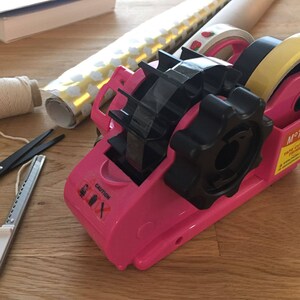 May include: A pink tape dispenser with a black handle and a black tape roll. The dispenser has a caution label with a warning symbol and the word "CAUTION".