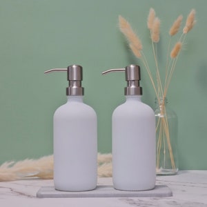 May include: Two white frosted glass soap dispensers with stainless steel pump tops. The dispensers sit on a gray and white marble tray. The background is a light green wall with dried decorative plants.