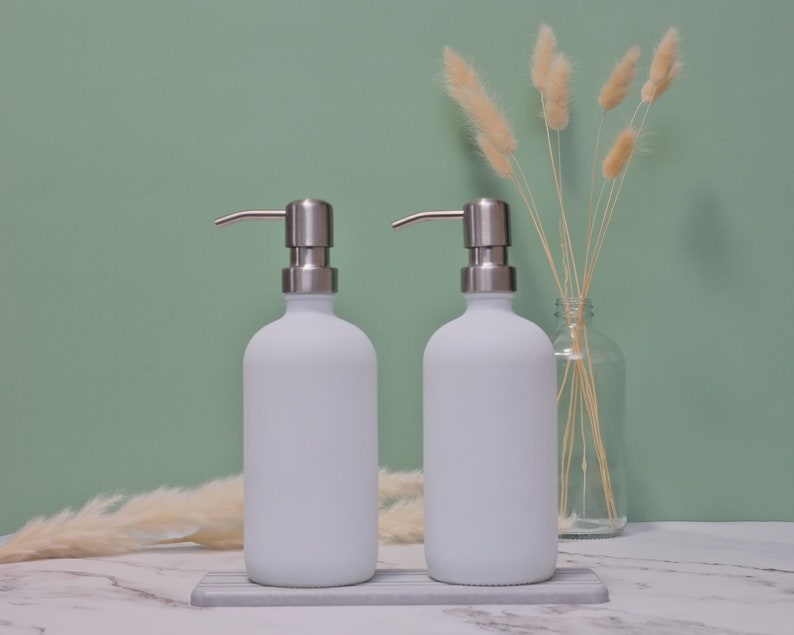 May include: Two white frosted glass soap dispensers with stainless steel pump tops. The dispensers sit on a gray and white marble tray. The background is a light green wall with dried decorative plants.