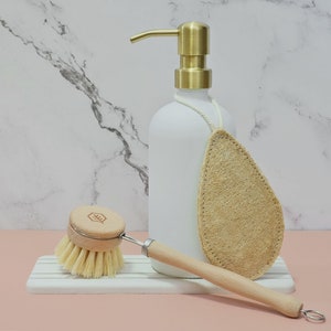 May include: A white soap dispenser with a gold pump, a wooden-handled dish brush, and a natural loofah sponge. The brush has a wooden head and light-colored bristles. The items are arranged on a white and gray striped tray.