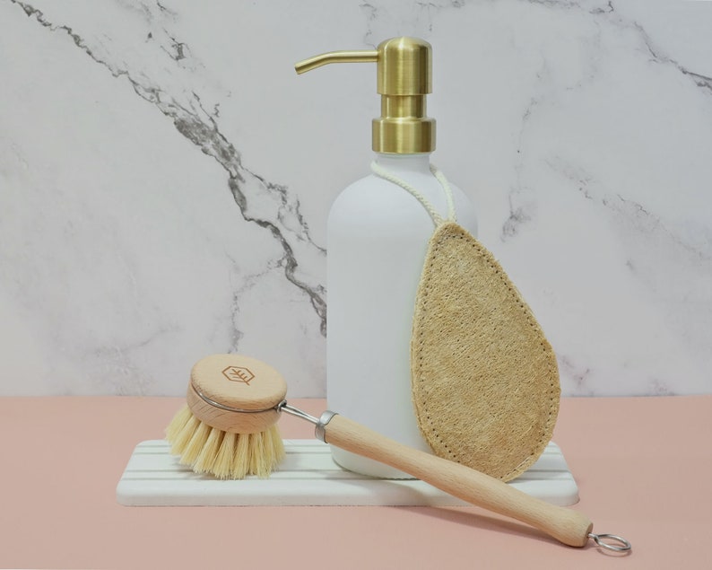 May include: A white soap dispenser with a gold pump, a wooden-handled dish brush, and a natural loofah sponge. The brush has a wooden head and light-colored bristles. The items are arranged on a white and gray striped tray.