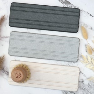 May include: Three rectangular dish scrub holders in gray, light gray, and off-white. The holders have horizontal grooves. A wooden dish brush with a leaf logo is on the off-white holder. The items are on a marble surface.