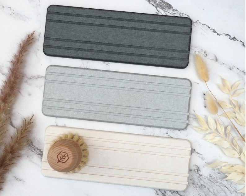 May include: Three rectangular dish scrub holders in gray, light gray, and off-white. The holders have horizontal grooves. A wooden dish brush with a leaf logo is on the off-white holder. The items are on a marble surface.