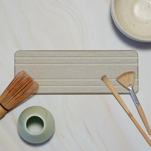 May include: A collection of matcha tea ceremony tools, including a bamboo whisk, a ceramic bowl, a brush, a scoop, and a small green ceramic bowl. The items are arranged on a light gray wooden tray, set against a white marble background.