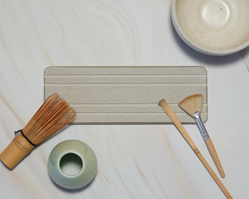 May include: A collection of matcha tea ceremony tools, including a bamboo whisk, a ceramic bowl, a brush, a scoop, and a small green ceramic bowl. The items are arranged on a light gray wooden tray, set against a white marble background.