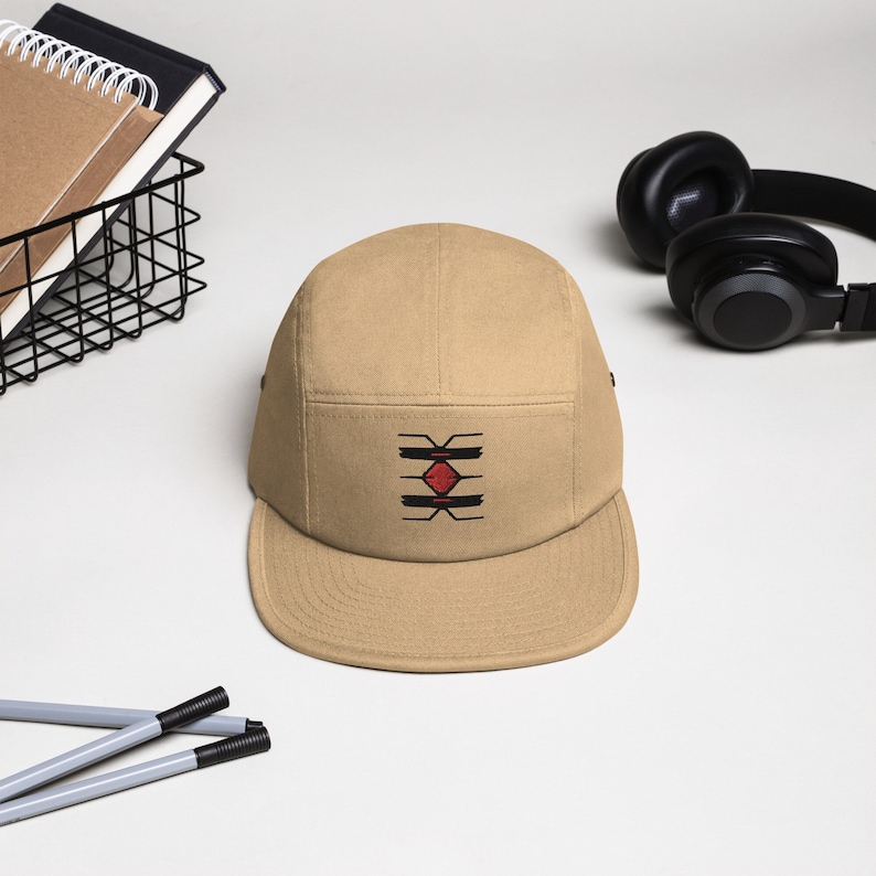 May include: A tan 5-panel cap with a black and red geometric design embroidered on the front.