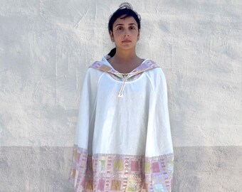 Könnte beinhalten: Ein weißer Kapuzenponcho mit einem Kordelzug am Ausschnitt und metallisch goldenen Akzenten. Der Saum und die Kapuze sind mit einem wiederkehrenden Muster aus quadratischen Aufnähern in Rosa, Gold und Hellgrün verziert, mit schimmerndem Rand.