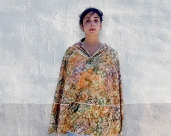 Könnte beinhalten: Ein Kapuzenponcho mit einem mehrfarbigen Blumenmuster in Orange-, Grün- und Brauntönen mit weißen, liniengezeichneten botanischen Details. Das Kleidungsstück hat eine lockere, drapierte Silhouette, einen V-Ausschnitt und wird über einer blauen Jeans getragen.
