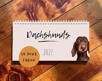 May include: A horizontal wall calendar with a light pink cover featuring the text Dachshunds and 2027 in black. A brown dachshund illustration is on the right side, and an orange circle with the text UK Print Edition is on the bottom left.