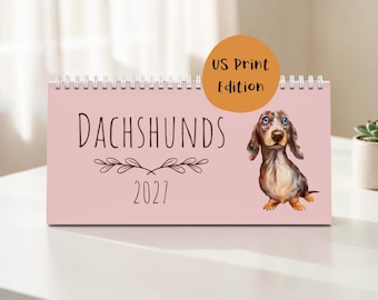 May include: A horizontal spiral-bound desk calendar with a light pink cover. The text reads "DACHSHUNDS 2027" in dark brown, accompanied by a brown and white illustration of a dachshund and an orange circular badge stating "US Print Edition".