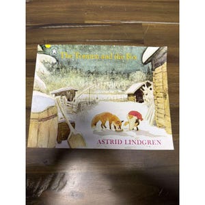 Può includere: Un libro per bambini intitolato "Il Tomten e la Volpe" di Astrid Lindgren. La copertina presenta una scena invernale con una volpe, uno gnomo e un paesaggio innevato. Il libro è su una superficie di legno.