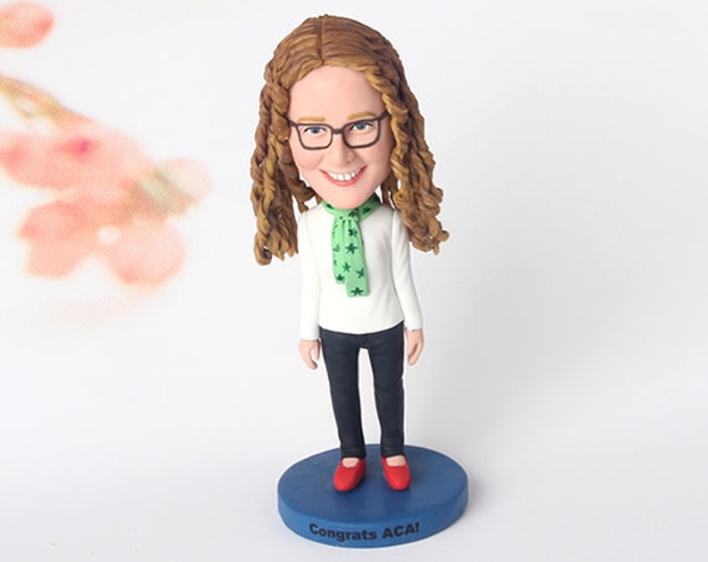 Peut inclure: Une figurine de t&ecirc;te de billard personnalis&eacute;e repr&eacute;sentant une personne aux cheveux bruns portant des lunettes, une chemise blanche, une &eacute;charpe verte &agrave; &eacute;toiles blanches, un pantalon noir et des chaussures rouges. La figurine est pos&eacute;e sur un socle bleu avec le texte "Congrats ACA!" imprim&eacute; dessus.