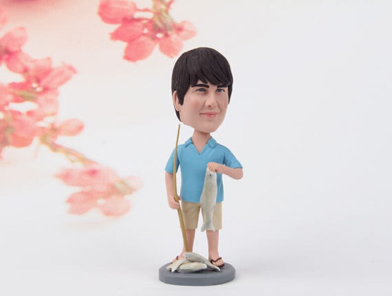 Puede incluir: Una figura personalizada de una persona sosteniendo una ca&ntilde;a de pescar y peces. La figura lleva una camisa azul y pantalones cortos color caqui. La figura est&aacute; de pie sobre una base gris.