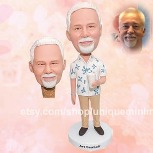 May include: A custom bobblehead figurine of a man with white hair and a goatee, wearing a white shirt with blue floral patterns, beige trousers, and brown shoes. The figurine holds a white cup. A second bobblehead head and a photo are also shown.