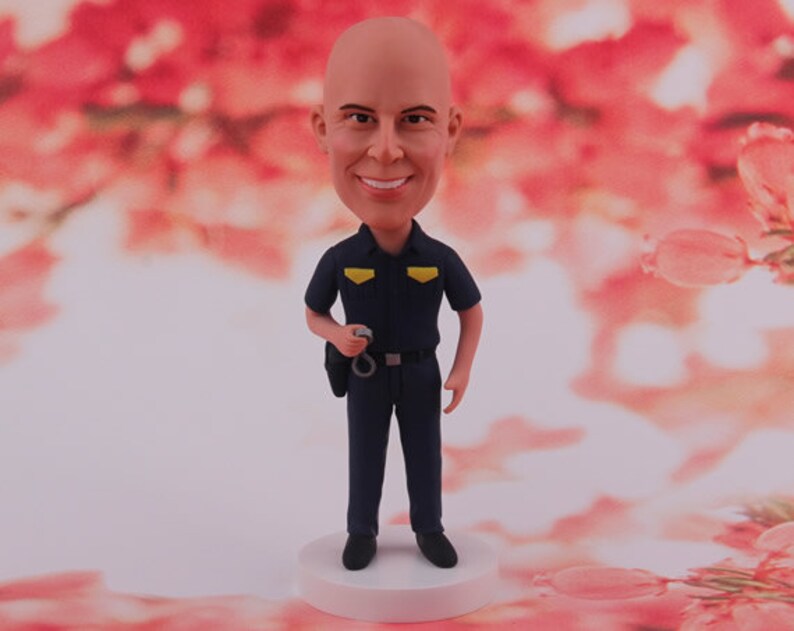 May include: A custom bobblehead figurine of a person wearing a blue police uniform with a yellow badge. The figurine is holding handcuffs in one hand and has a serious expression on their face.