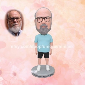 May include: A custom bobblehead figurine of a person with a grey beard and glasses wearing a light blue t-shirt and black shorts. The bobblehead is standing on a round base with the text "Dr. Charles Conrad, DGS" printed on it.