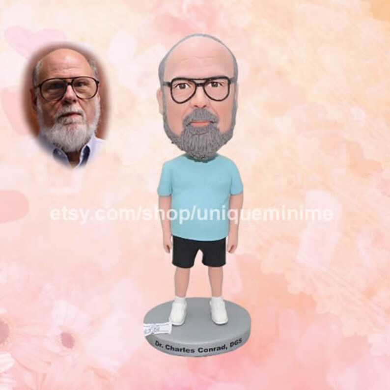 May include: A custom bobblehead figurine of a person with a gray beard and glasses wearing a light blue t-shirt and black shorts. The bobblehead is standing on a round base with the text "Dr. Charles Conrad, DGS" printed on it.