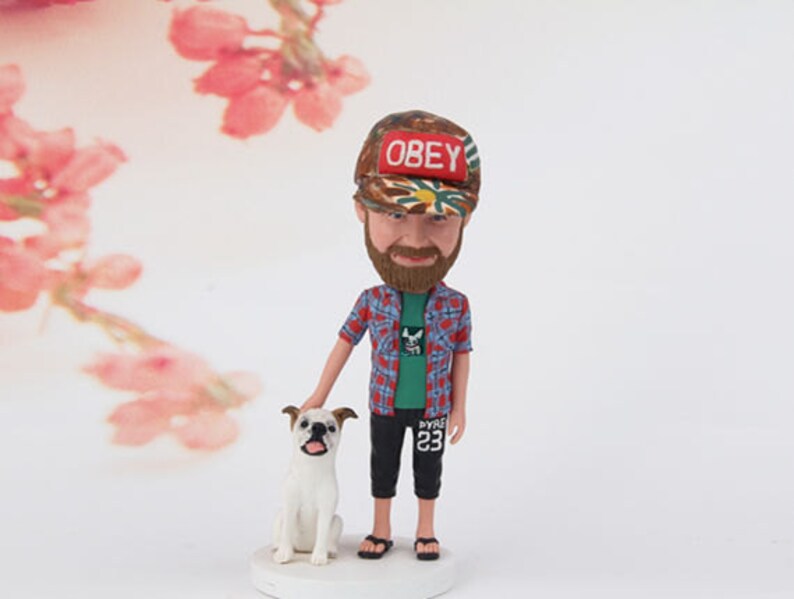 May include: A custom bobblehead figurine of a person wearing a camouflage baseball cap with the word "OBEY" on it, a green and black plaid shirt, green t-shirt, black pants, and a beard. The figurine is holding a white dog with brown spots.
