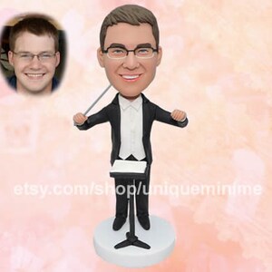 May include: A custom figurine of a person in a black tuxedo holding a conductor's baton. The figurine is standing on a black stand with a music stand. The person is wearing glasses and has a white shirt with a black bow tie. The figurine is on a white background.